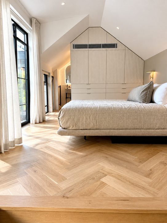 An attic bedroom with downlights, a brass wall light above the bed and a lit ensuite beyond the wardrobe