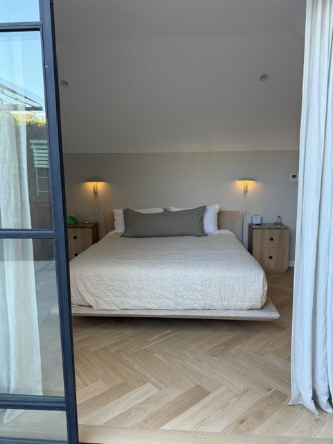 A bedroom seen through a steel-framed door, with a wall light above each bedside table