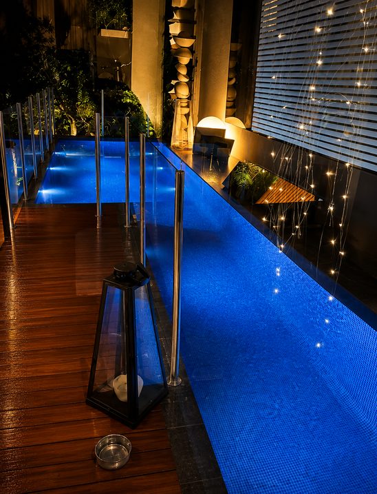 A narrow lap pool lit blue at night beside a timber deck, with a glass fence, fairy lights and a lantern