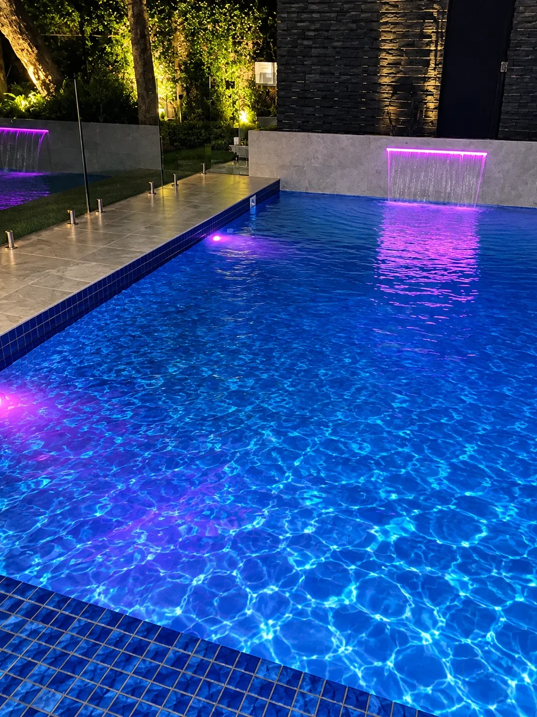 A pool lit at night, with magenta-lit water blades and garden uplights behind a glass fence