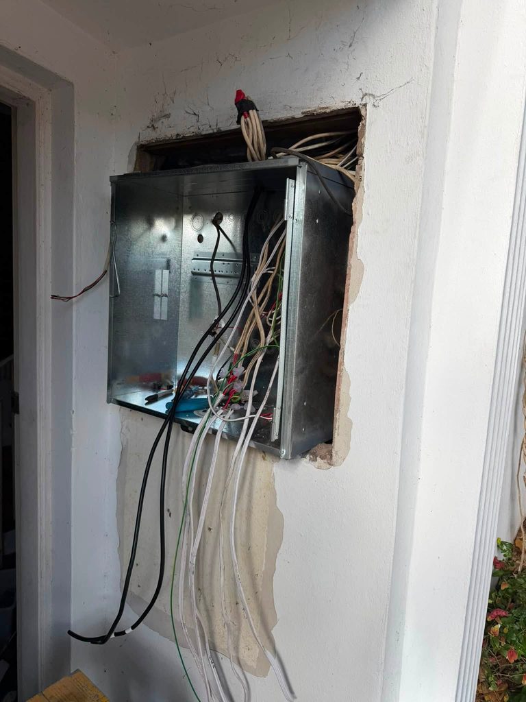 An old switchboard enclosure opened up in a wall, with cables hanging from it