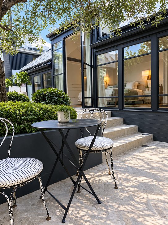 A courtyard with a bistro table, stone steps with in-ground lights, and a bedroom lit by wall lights behind steel-framed doors
