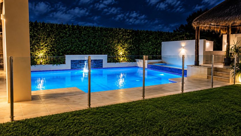 A pool lit blue at night behind a glass fence, with hedge uplights and a lit water feature