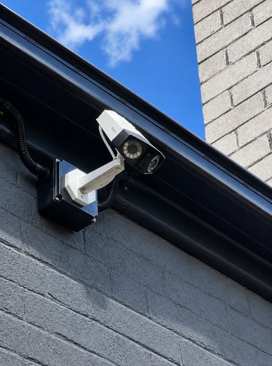 A security camera on a white bracket under a dark eave, its cable run in conduit