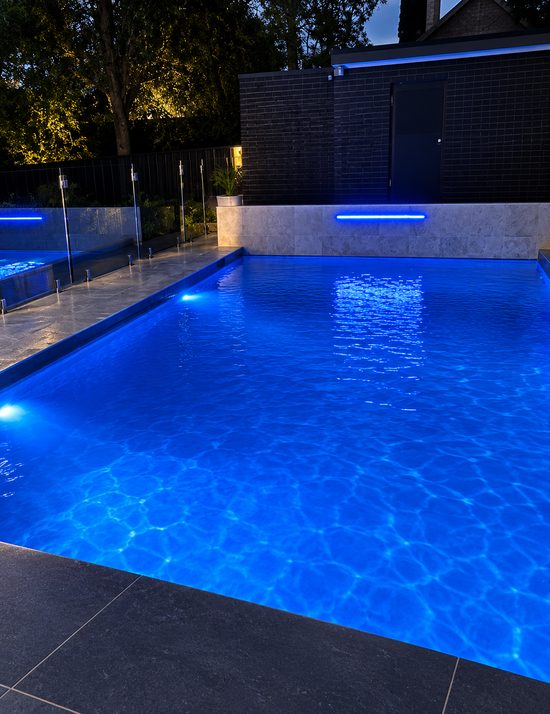 A pool lit blue from underwater at dusk, with a glass fence and a blue light strip on the wall